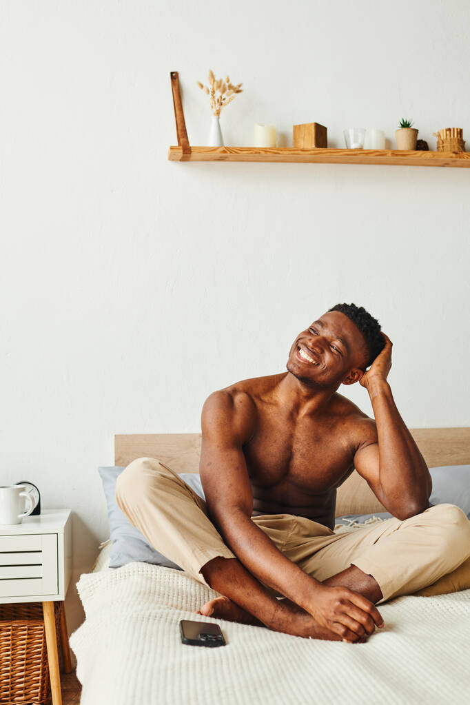 笑うシャツのないアフリカ系アメリカ人の男 パジャマパンツ 自宅でベッドでスマートフォンの近くを探して - 写真・画像
