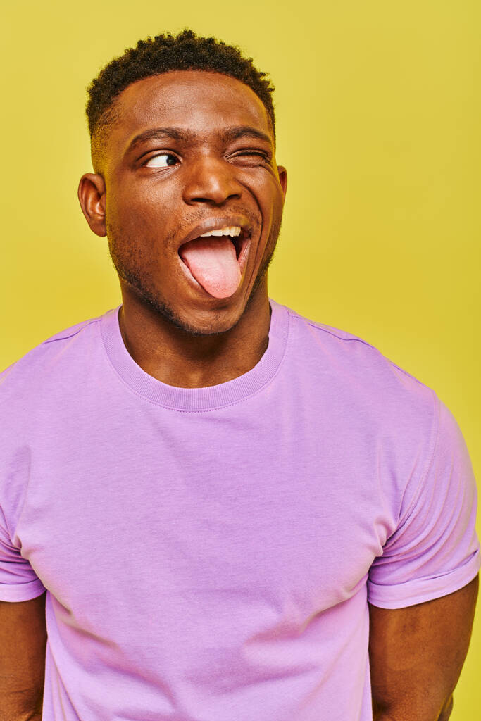 divertido afroamericano hombre en púrpura camiseta sobresaliendo lengua y mirando hacia otro lado en amarillo telón de fondo - Foto, Imagen