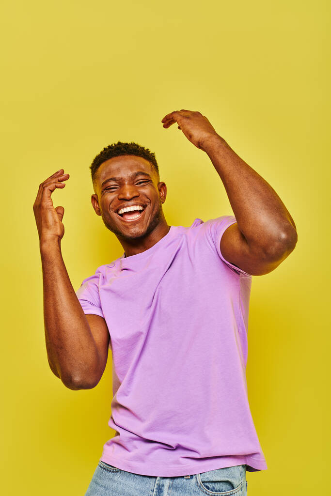 podekscytowany i modny Afrykański Amerykanin w fioletowej koszulce gestykulujący śmiejąc się na żółtym tle - Zdjęcie, obraz