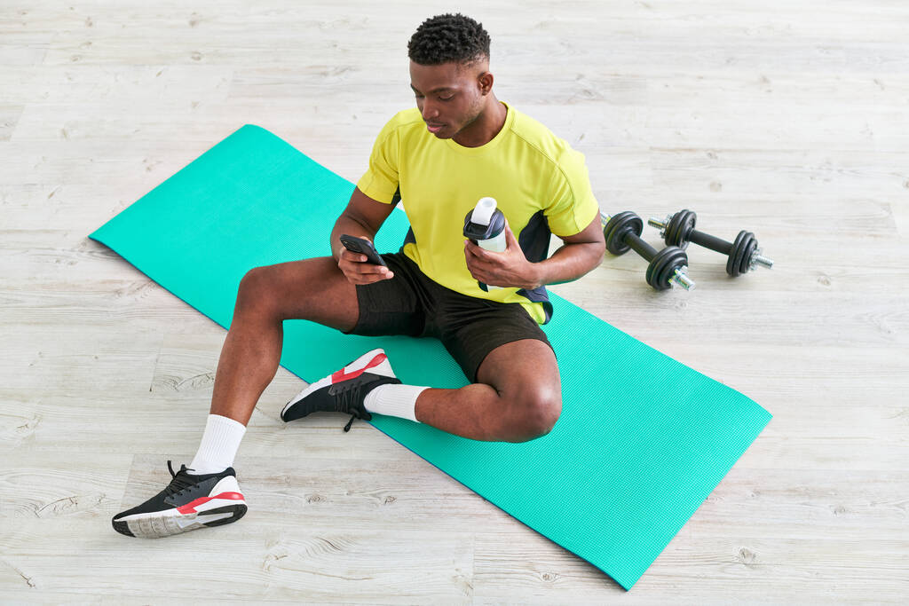 fitt afro-amerikai férfi sportruházat ül fitness mat okostelefon és sport palack - Fotó, kép