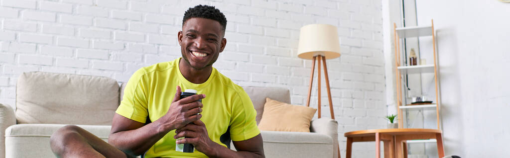 gelukkige sportieve Afrikaan amerikaanse man met sportfles kijken naar camera in moderne woonkamer, banner - Foto, afbeelding