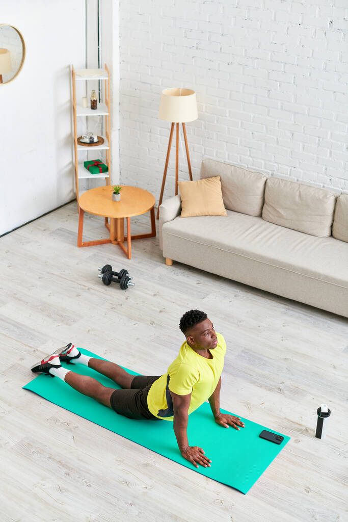 Spor kıyafetli Afro-Amerikan bir adamın spor kıyafetleri içinde spor minderinde yılan yoga pozu verirken yüksek açılı görüntüsü. - Fotoğraf, Görsel