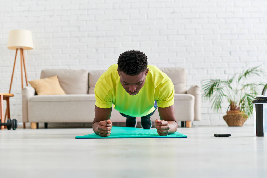 Afrikanisch-amerikanischer Mann im Sportbekleidungstraining in Plank-Pose auf Fitnessmatte im modernen Wohnzimmer - Foto, Bild