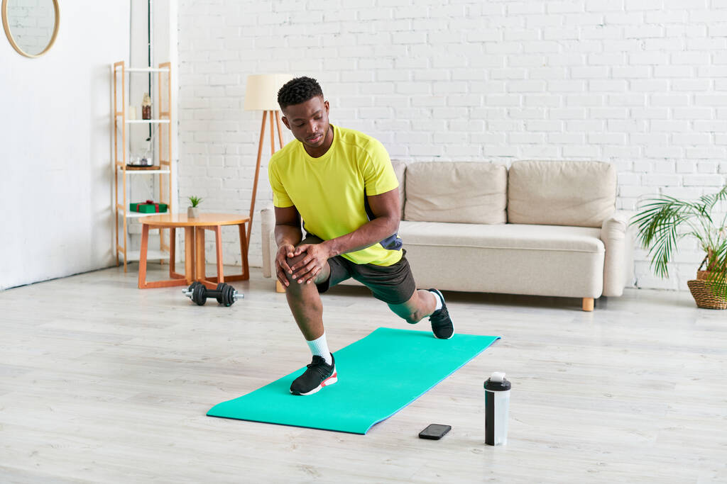 sportovní africký Američan chlap pracuje na fitness mat v blízkosti sportovní láhve a smartphone doma - Fotografie, Obrázek