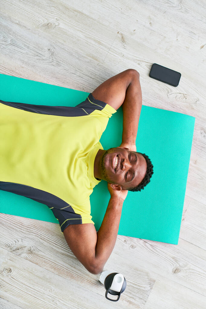 Blick von oben auf einen Afroamerikaner in Sportbekleidung, der sich mit geschlossenen Augen auf eine Fitnessmatte legt - Foto, Bild