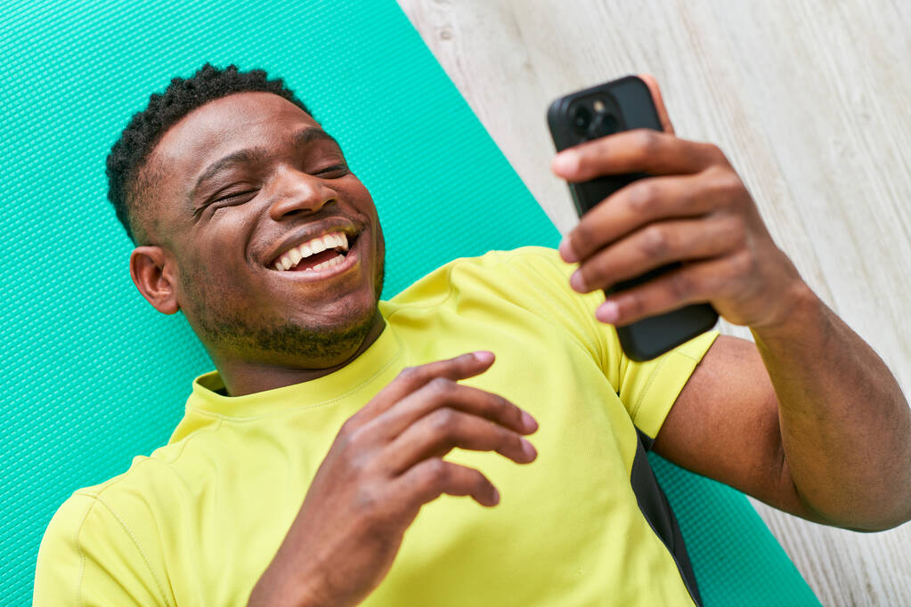 junger fröhlicher afrikanisch-amerikanischer Mann in Sportbekleidung surft auf Smartphone im Internet, Draufsicht - Foto, Bild