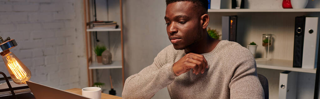 νέος συγκεντρωμένος Αφροαμερικάνος ελεύθερος επαγγελματίας που εργάζεται στο lap-top το βράδυ στο σπίτι, banner - Φωτογραφία, εικόνα
