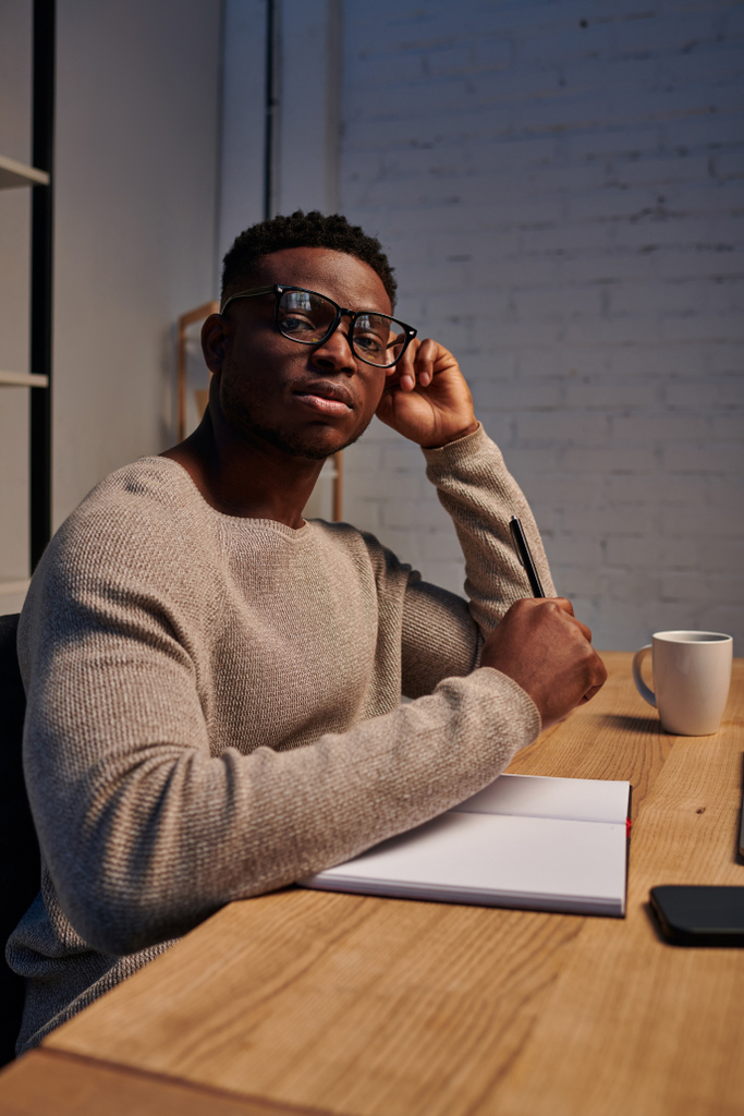 афроамериканський чоловік в окулярах дивиться на камеру біля записника в домашньому офісі, фрілансер - Фото, зображення