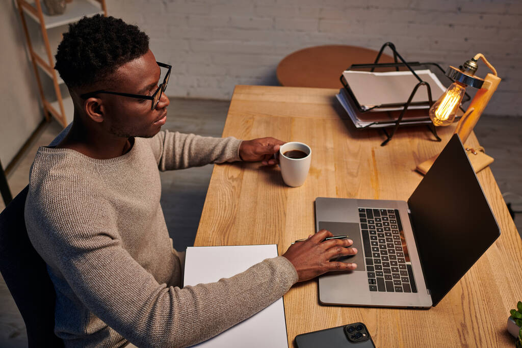 homem americano africano em óculos segurando xícara de café e trabalhando no laptop no escritório em casa à noite - Foto, Imagem