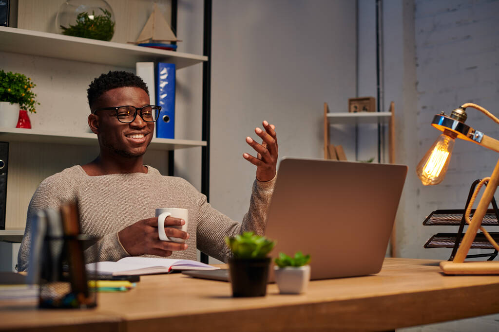 Kahve fincanı taşıyan mutlu Afro-Amerikalı adam video odasında el kol hareketi yapıyor, serbest çalışan. - Fotoğraf, Görsel