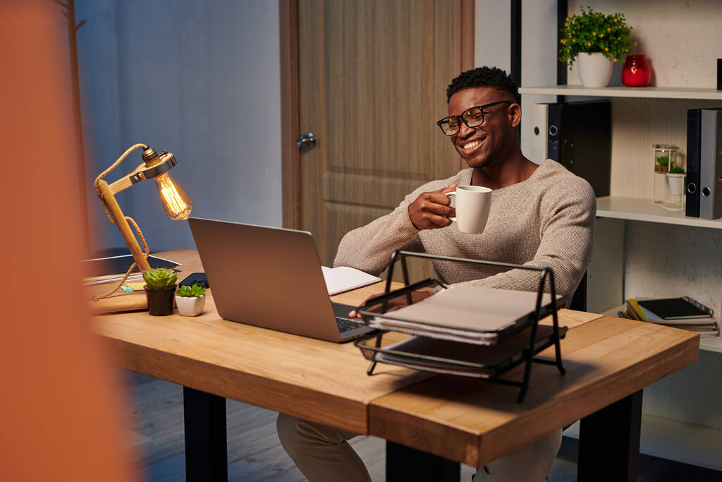 homem americano africano alegre com copo de café sorrindo durante chamada de vídeo em home office, freelancer - Foto, Imagem
