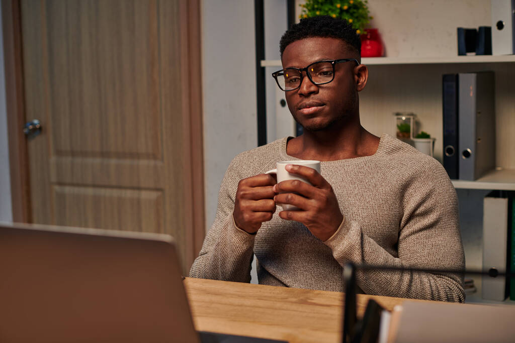 hombre afroamericano reflexivo con taza de café mirando el ordenador portátil mientras trabaja en casa por la noche - Foto, Imagen