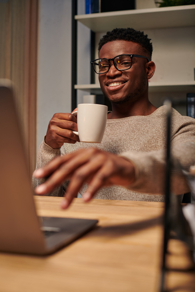 joyeux freelance afro-américain avec tasse de café travaillant à l'ordinateur portable dans le bureau à la maison la nuit - Photo, image