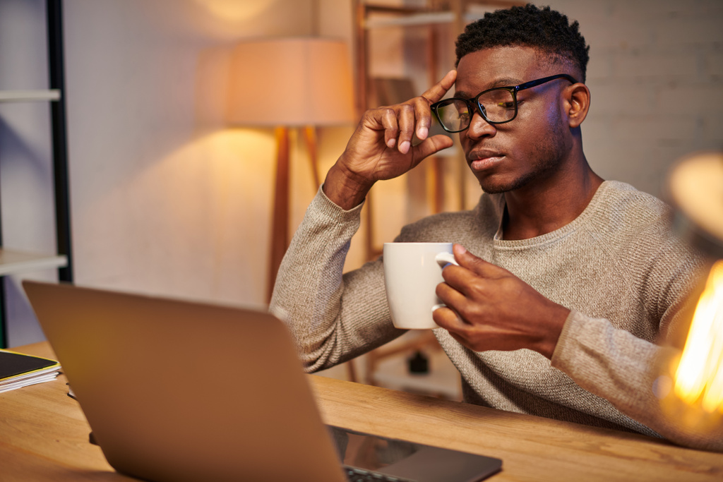 pensativo Africano americano freelancer com xícara de café sentado perto de laptop à noite em casa escritório - Foto, Imagem