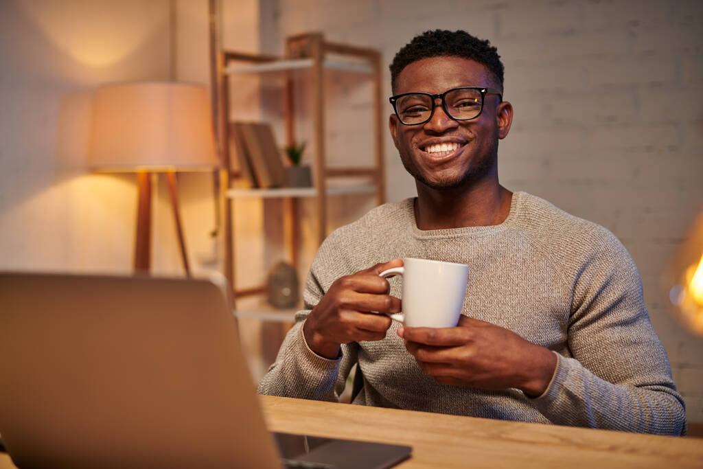веселий афроамериканський фрілансер з чашкою кави сидить біля ноутбука вночі в домашньому офісі - Фото, зображення