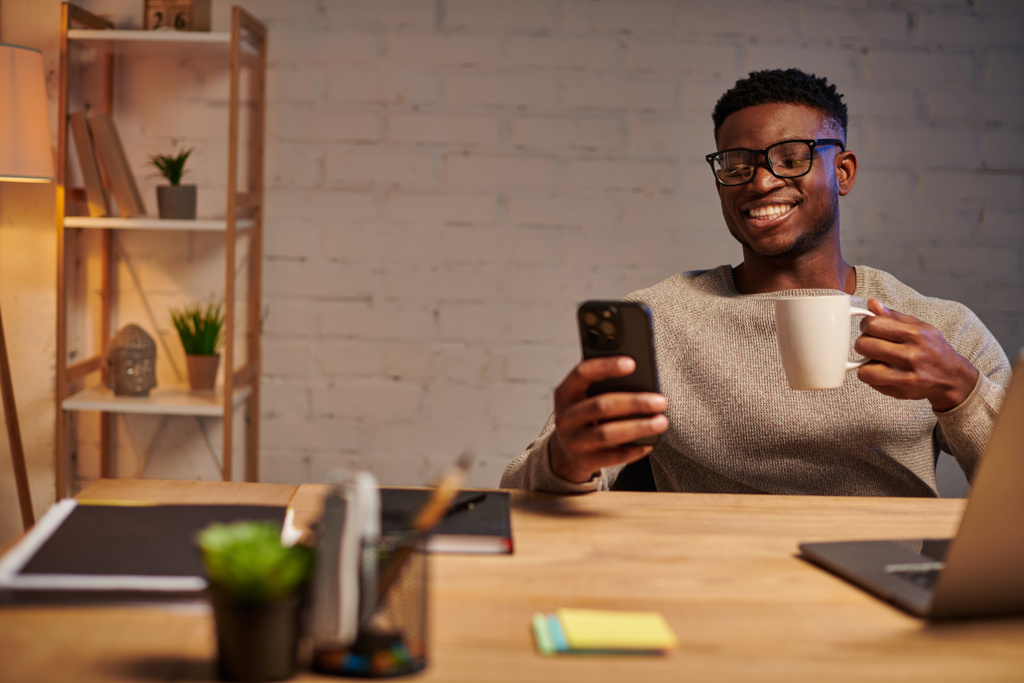 幸せなアフリカ系アメリカ人フリーランサーとコーヒーカップとスマートフォン 夜の自宅オフィスでラップトップに近い - 写真・画像