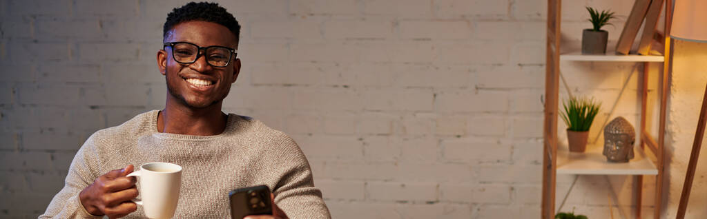 freelance afroamericano sorridente con tazza di caffè e smartphone che lavora in ufficio, banner - Foto, immagini