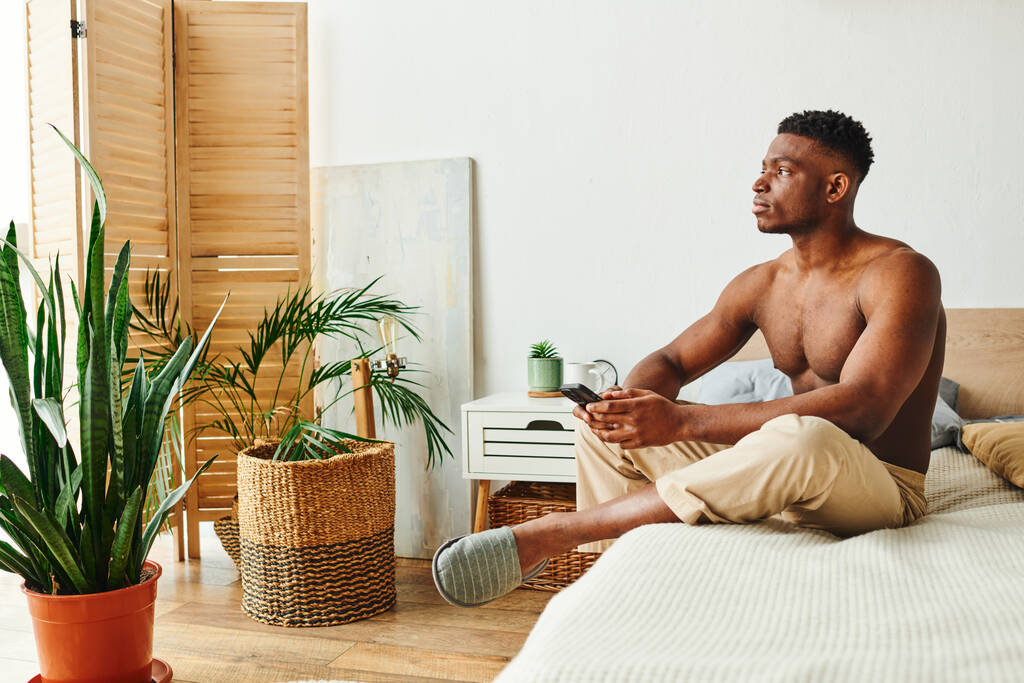 hombre afroamericano reflexivo con teléfono inteligente sentado y mirando hacia otro lado en el dormitorio con decoración verde - Foto, Imagen