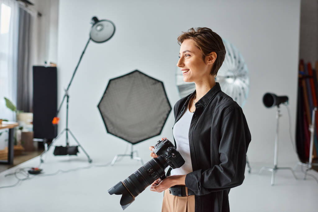 alegre fotógrafa femenina en traje casual con cámara en sus manos sonriendo y mirando hacia otro lado - Foto, Imagen