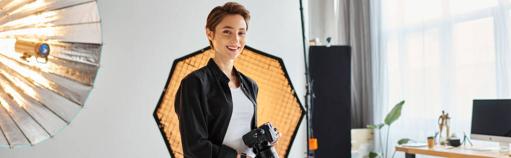 привлекательная веселая женщина в повседневной одежде позирует в своей студии с камерой в руках, баннер - Фото, изображение