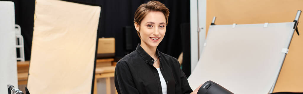 veselá krátkovlasá žena s fotografickou technikou, šťastně se usmívající na kameru ve svém studiu, banner - Fotografie, Obrázek
