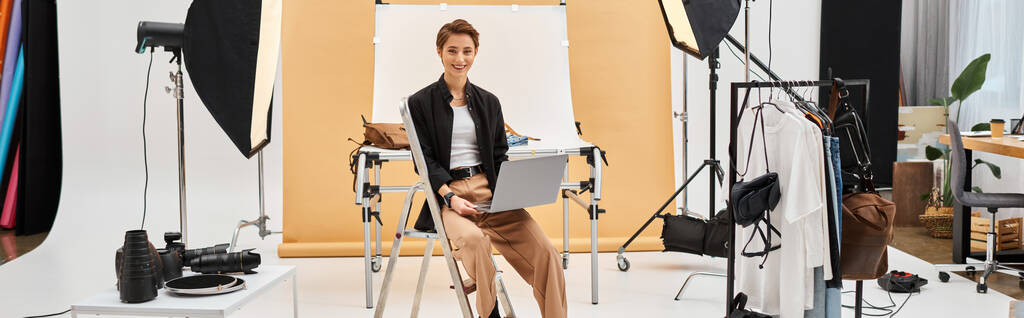 fröhliche Frau, die mit Laptop sitzt, während sie Fotos retuschiert und im Studio in die Kamera lächelt, Banner - Foto, Bild