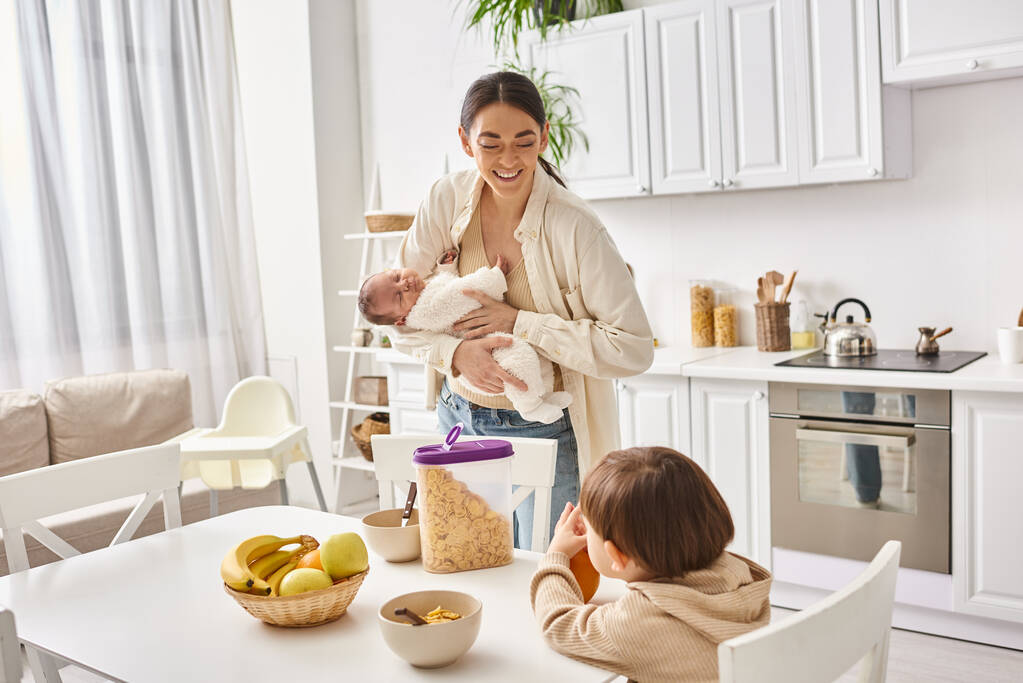 ευτυχισμένη ελκυστική μητέρα απολαμβάνοντας πρωινό με το παιδί και τα νεογέννητα γιους της, σύγχρονη ανατροφή των παιδιών - Φωτογραφία, εικόνα