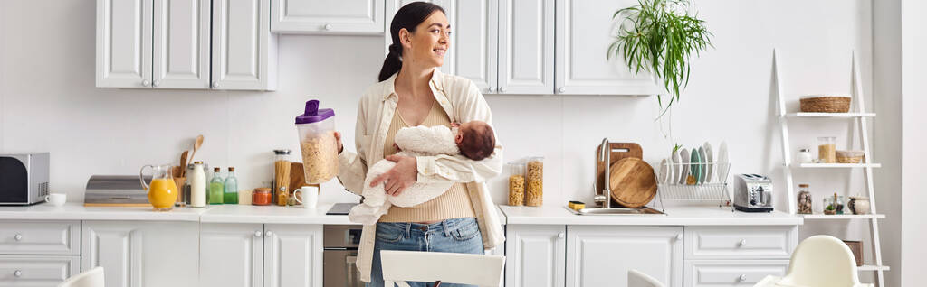 beautiful joyous woman in comfy homewear holding her cute newborn baby while preparing breakfast - Photo, Image