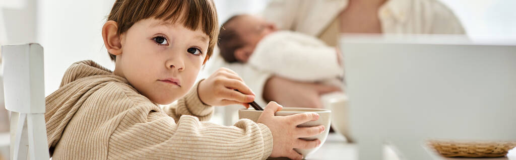 чарівний хлопчик дивиться на камеру під час сніданку зі своєю розмитою мамою і братом на фоні, банер - Фото, зображення