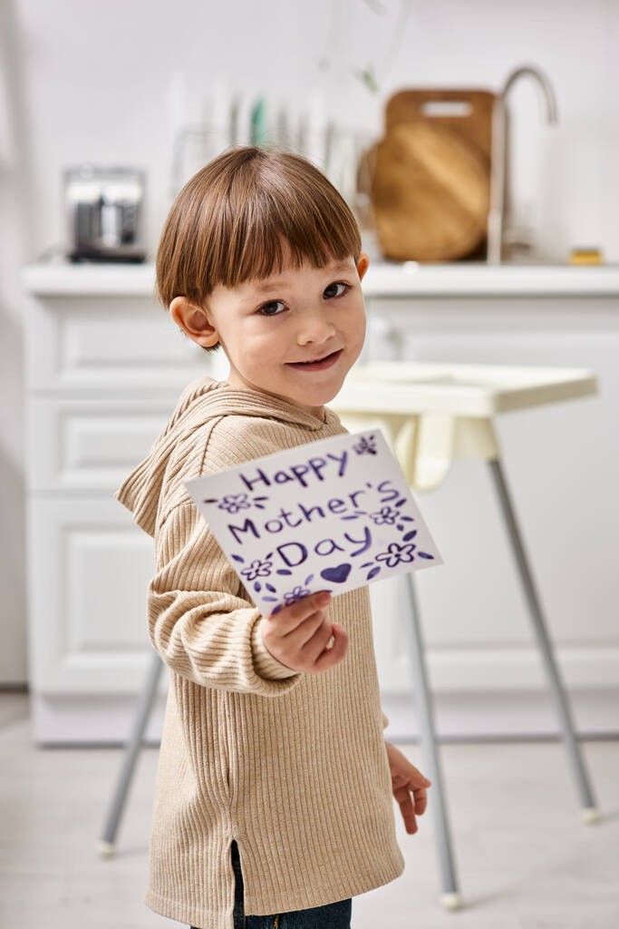 adorable cheerful toddler boy posing with greeting card for Mothers day and smiling at camera - Photo, Image