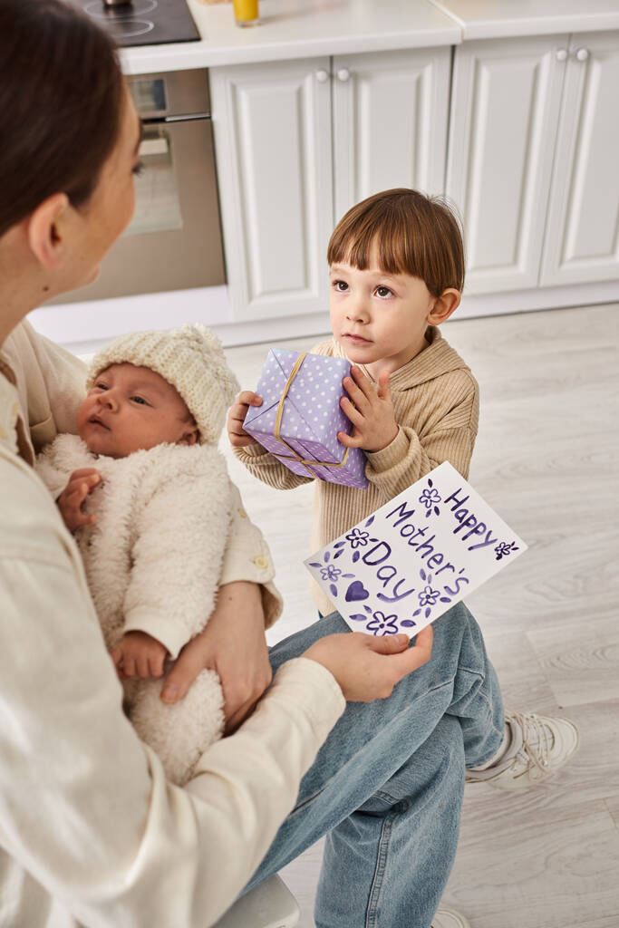 schattig jongen geven geschenk en ansichtkaart naar zijn moeder terwijl ze het houden van zijn pasgeboren broer, Moederdag - Foto, afbeelding