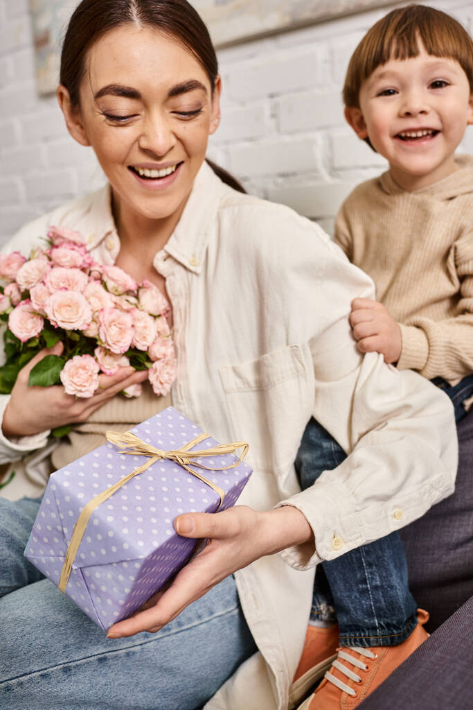 amando mãe feliz posando no sofá com seu filho criança com buquê de flores e presente, dia das mães - Foto, Imagem