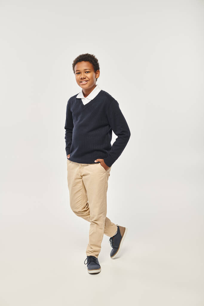onnellinen afrikkalainen amerikkalainen koulupoika siisti univormu seisoo kädet taskussa harmaalla taustalla - Valokuva, kuva