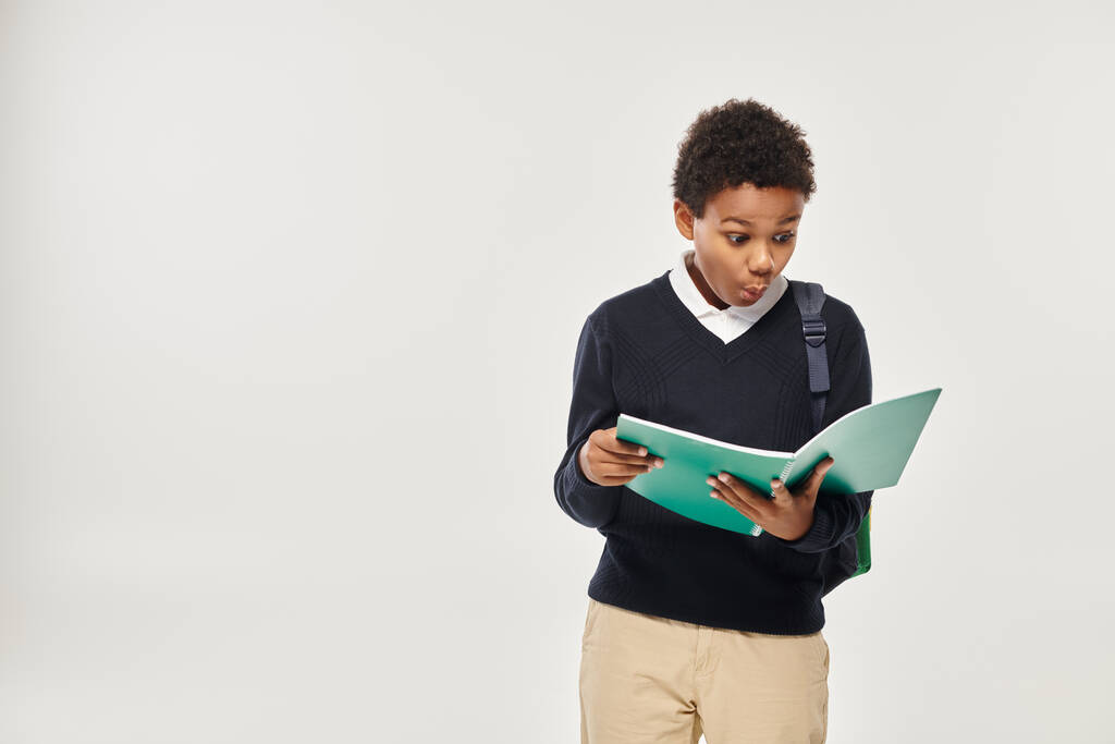 geschokt afrikaanse amerikaanse schooljongen in uniform kijken naar notities in het leerboek over grijze achtergrond - Foto, afbeelding