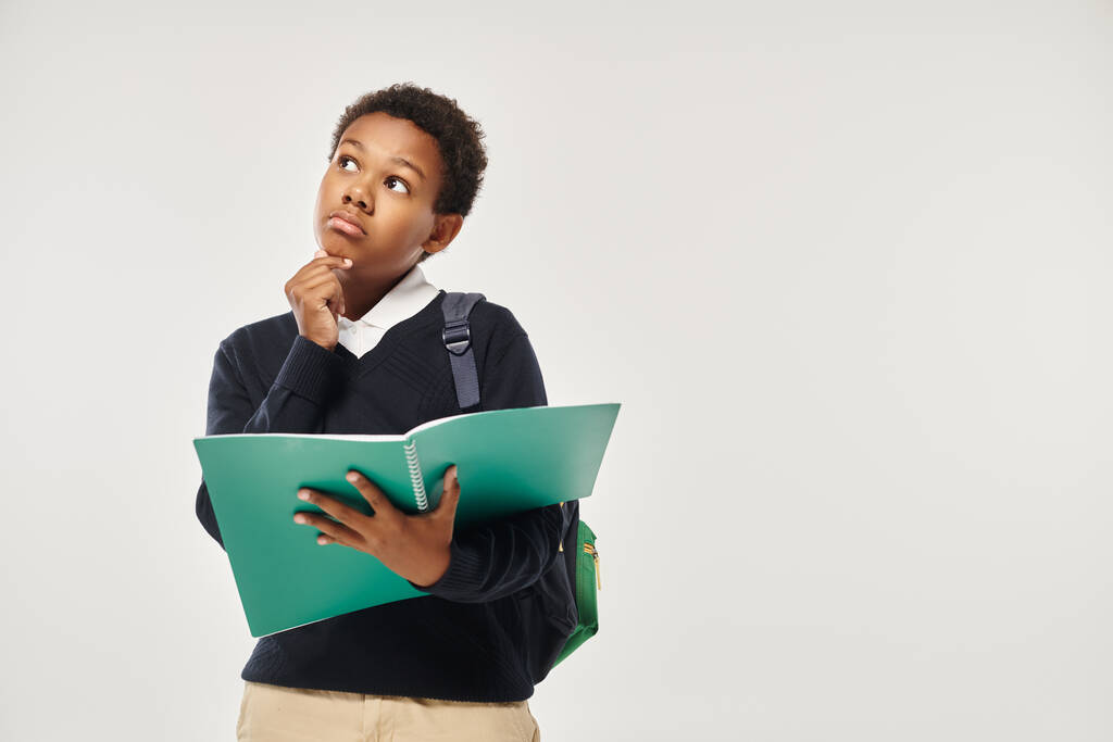 nachdenklicher afrikanisch-amerikanischer Schuljunge in Uniform mit Notizbüchern auf grauem Hintergrund - Foto, Bild