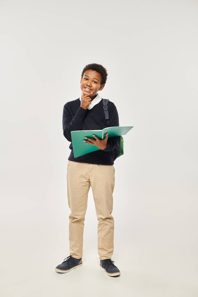 šťastný africký americký školák v uniformě držení notebook na šedém pozadí, ruka blízko brady - Fotografie, Obrázek