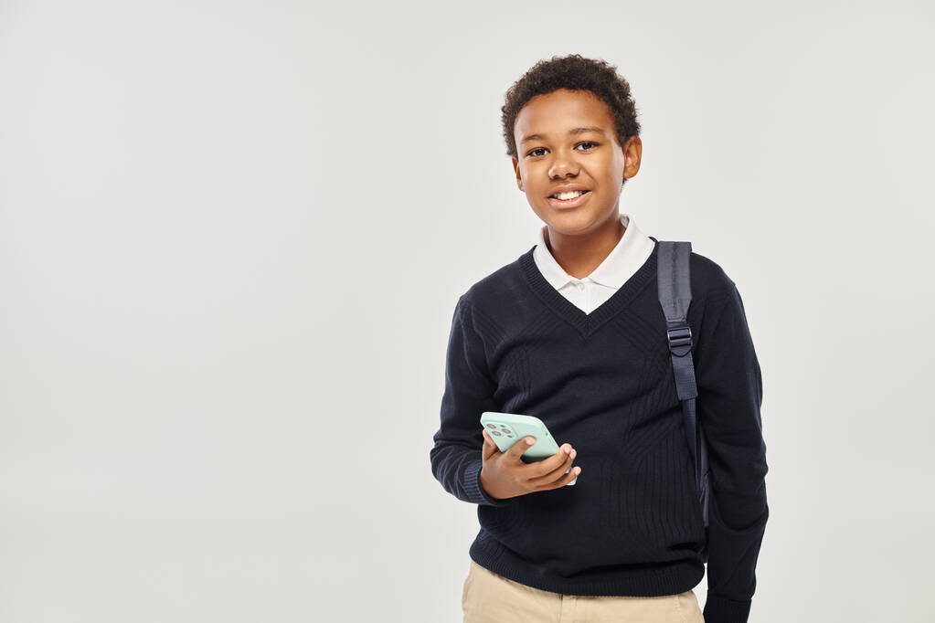 スマートフォンを持ち,灰色の背景に立っているユニフォームで幸せなアフリカ系アメリカ人のボーイ - 写真・画像
