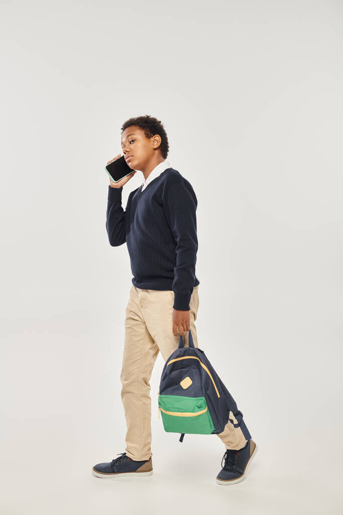 afrikanisch-amerikanischer Schüler in Uniform spricht mit Smartphone und geht mit Rucksack auf grau - Foto, Bild