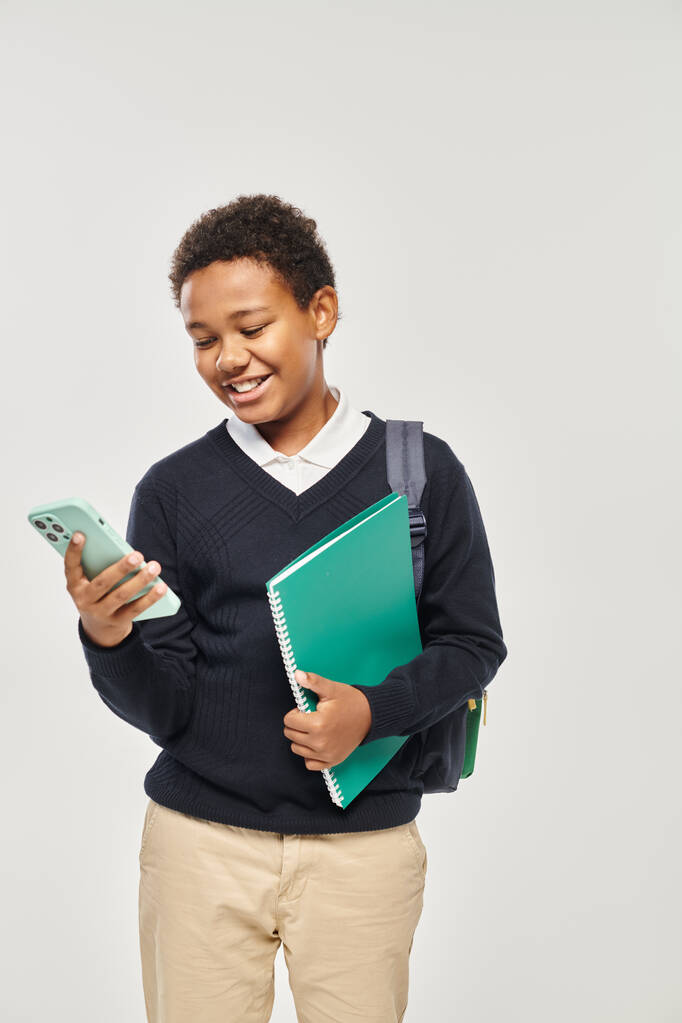 szczęśliwy Afrykański amerykański uczeń w mundurze trzymając smartfona i notatnik na szarym tle - Zdjęcie, obraz