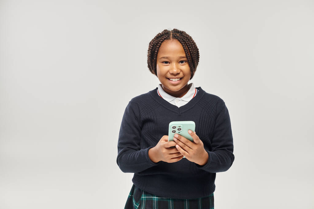 グレーの背景にスマートフォンを保持する制服の幸せなアフリカ系アメリカ人女子高生 - 写真・画像