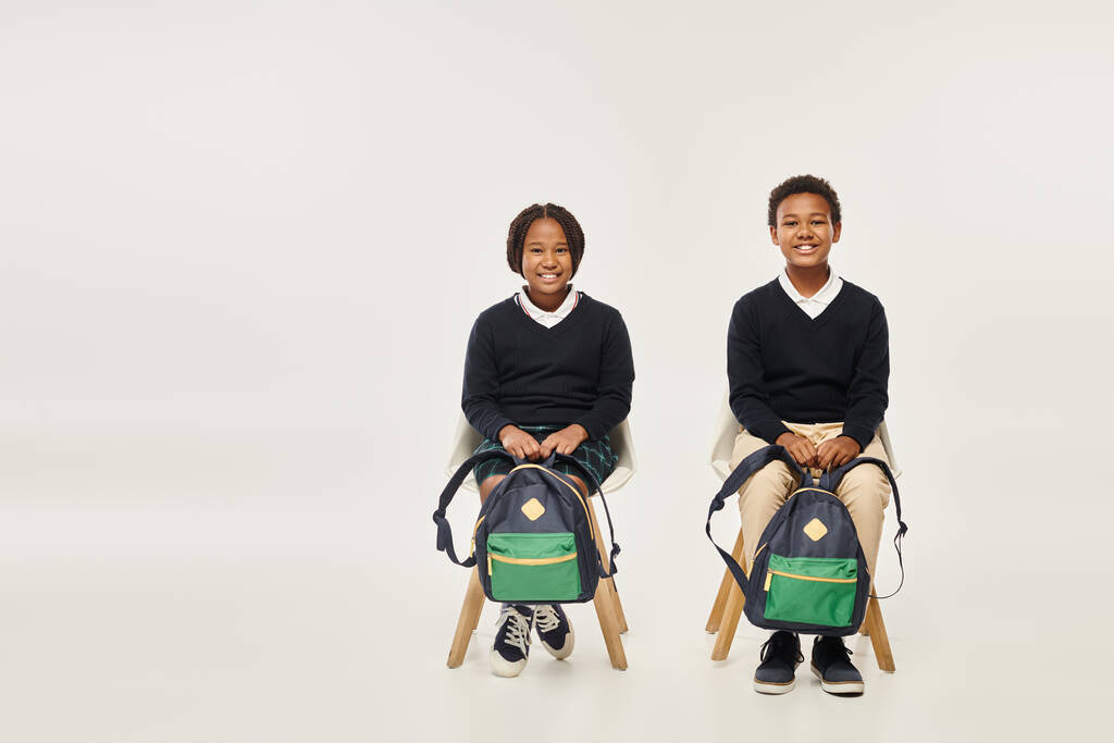 joyeux écoliers afro-américains avec des sacs à dos assis et regardant la caméra sur fond gris - Photo, image