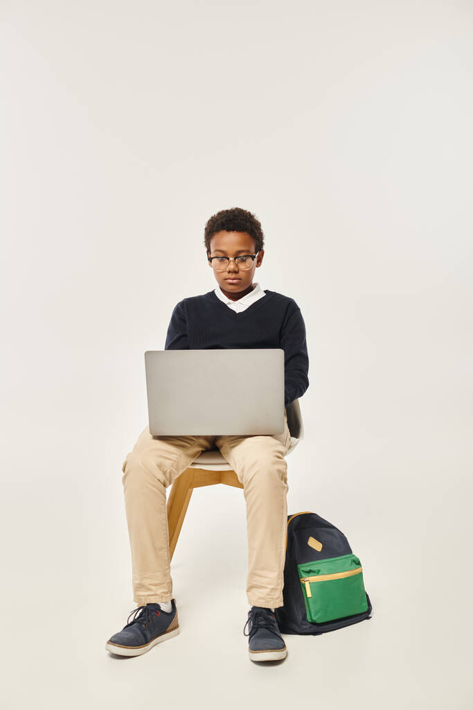 écolier afro-américain concentré en uniforme et lunettes à l'aide d'un ordinateur portable et assis sur fond gris - Photo, image