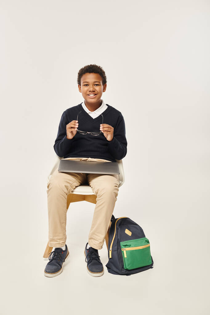 happy african american schoolboy in uniform holding glasses and sitting with laptop on grey backdrop - Photo, Image