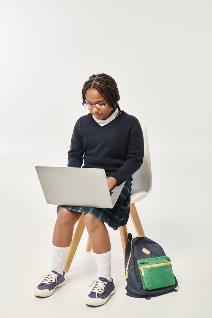 εστιασμένη Αφροαμερικανίδα μαθήτρια με στολή και γυαλιά που χρησιμοποιεί φορητό υπολογιστή και κάθεται σε γκρι φόντο - Φωτογραφία, εικόνα
