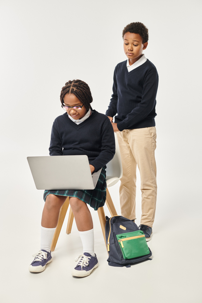 africano americano ragazzo in uniforme in piedi vicino eccitato studentessa utilizzando computer portatile su sfondo grigio - Foto, immagini