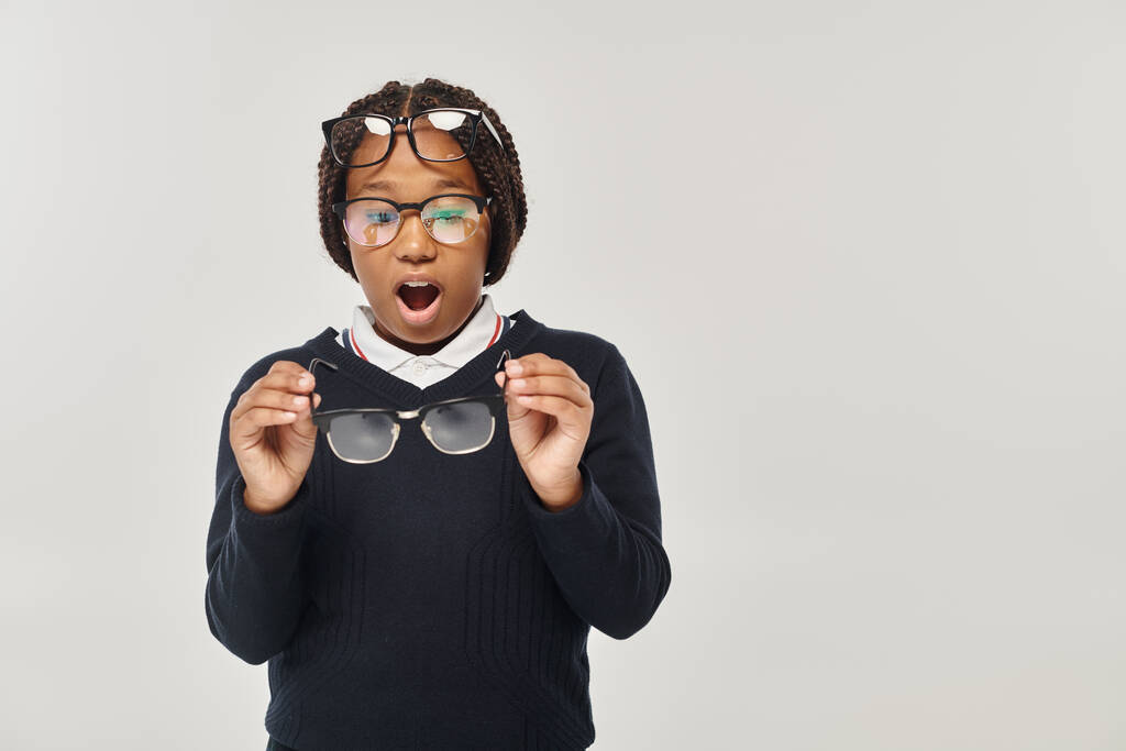 wstrząśnięta afrykańska dziewczyna w swetrze i okularach, trzymająca okulary i patrząca w kamerę na szarości - Zdjęcie, obraz