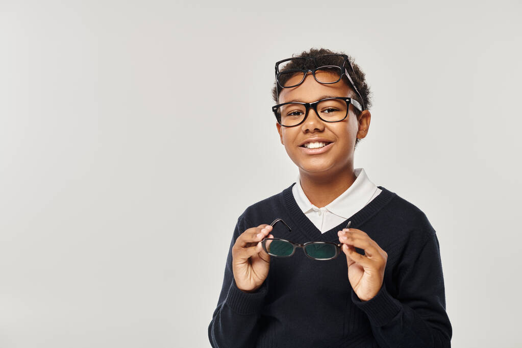 happy african american schoolboy in eyewear holding glasses and looking at camera on grey backdrop - Photo, Image