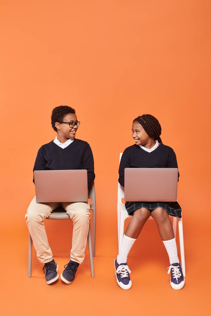 niños afroamericanos felices en uniforme sentados en sillas y usando computadoras portátiles sobre fondo naranja - Foto, Imagen