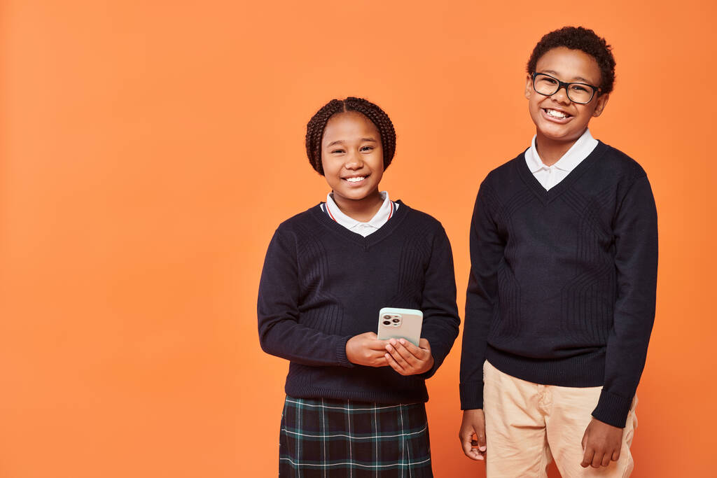 веселые африканские американские школьники в форме, смотрящие на смартфон на оранжевом фоне - Фото, изображение