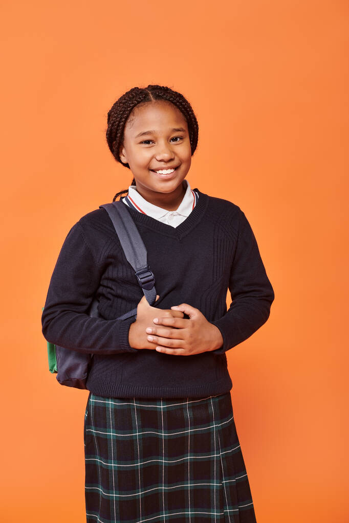 glücklich afrikanisch-amerikanische Schulmädchen in Uniform lächelnd und mit Rucksack auf orangefarbenem Hintergrund - Foto, Bild